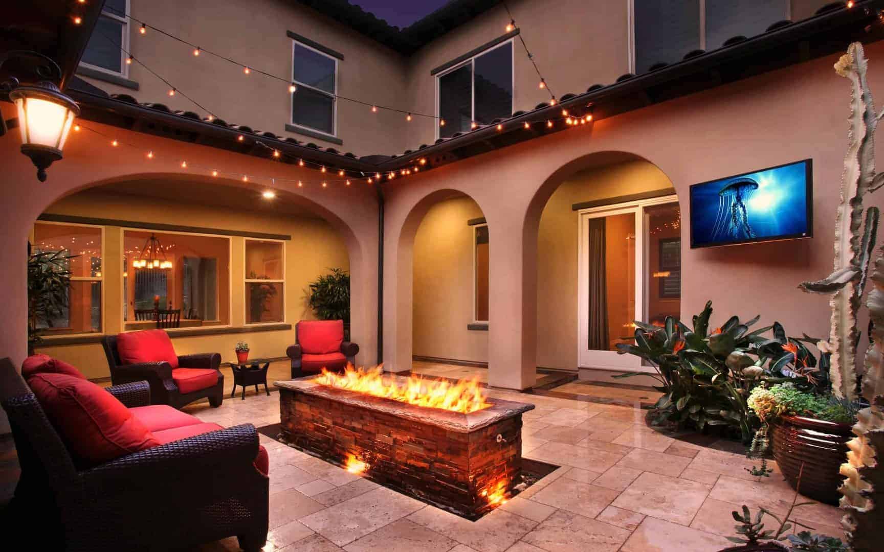 Cozy nighttime patio with stone fire pit, red chairs, string lights, and mounted TV showing jellyfish